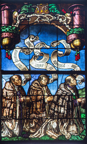 Three brothers from the Suter family are shown praying in front of three patron saints of the family. The hilly meadow landscape in the background running through three of the stained glass windows, the deep blue sky and the colourful Renaissance ornamentation with decorative branches in the form of arches suggest that the stained glass windows are based on designs by Hans Baldung Grien.